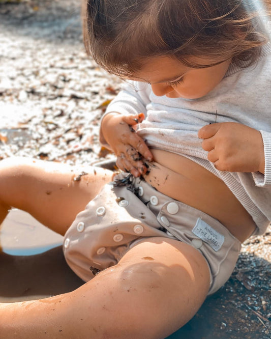 Toddler sits rubbing mud on her belly wearing a tan modern cloth nappy and grey shirt