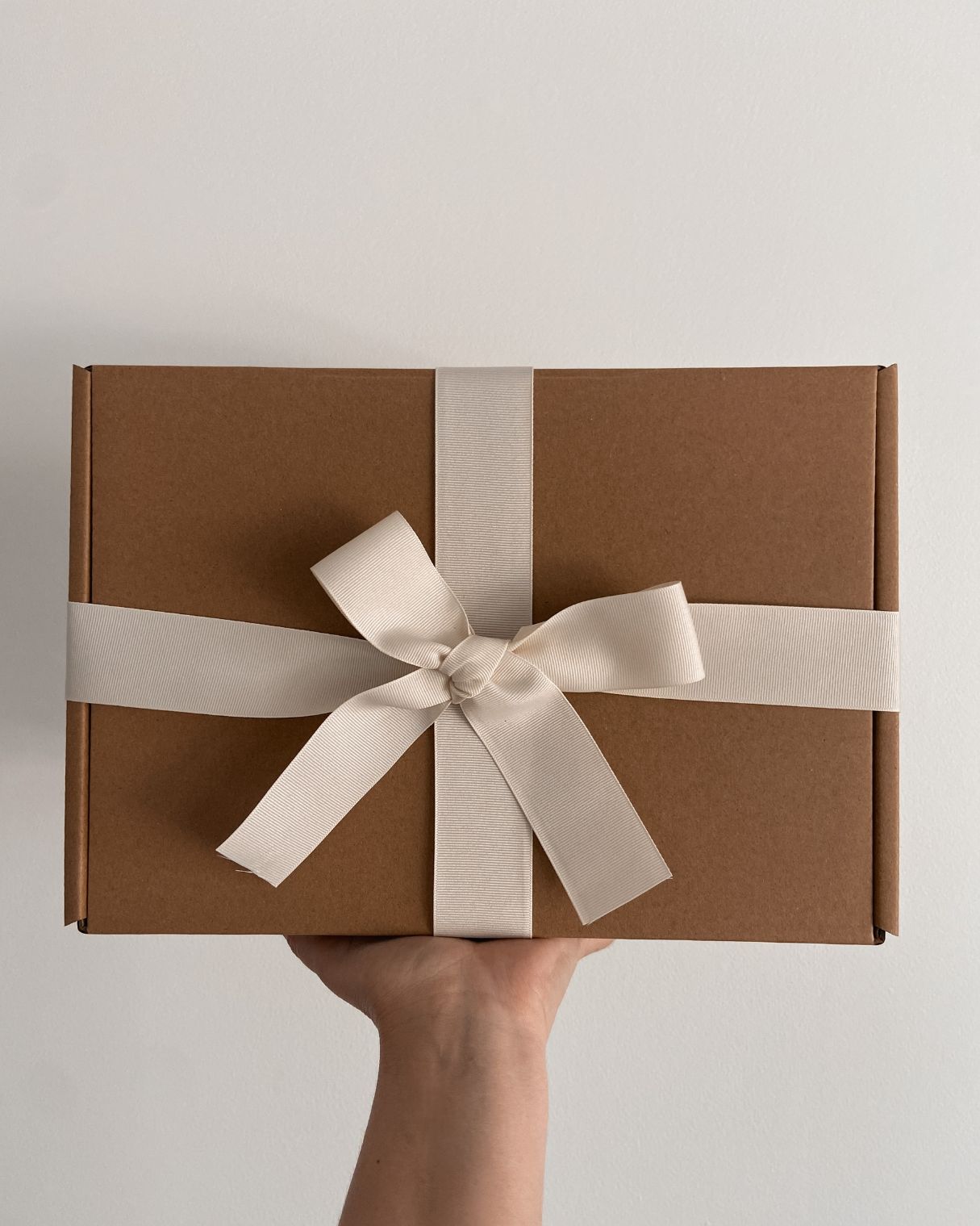 Gift wrap box being held high. Box is natural cardboard with a thick cream ribbon tied in a bow.