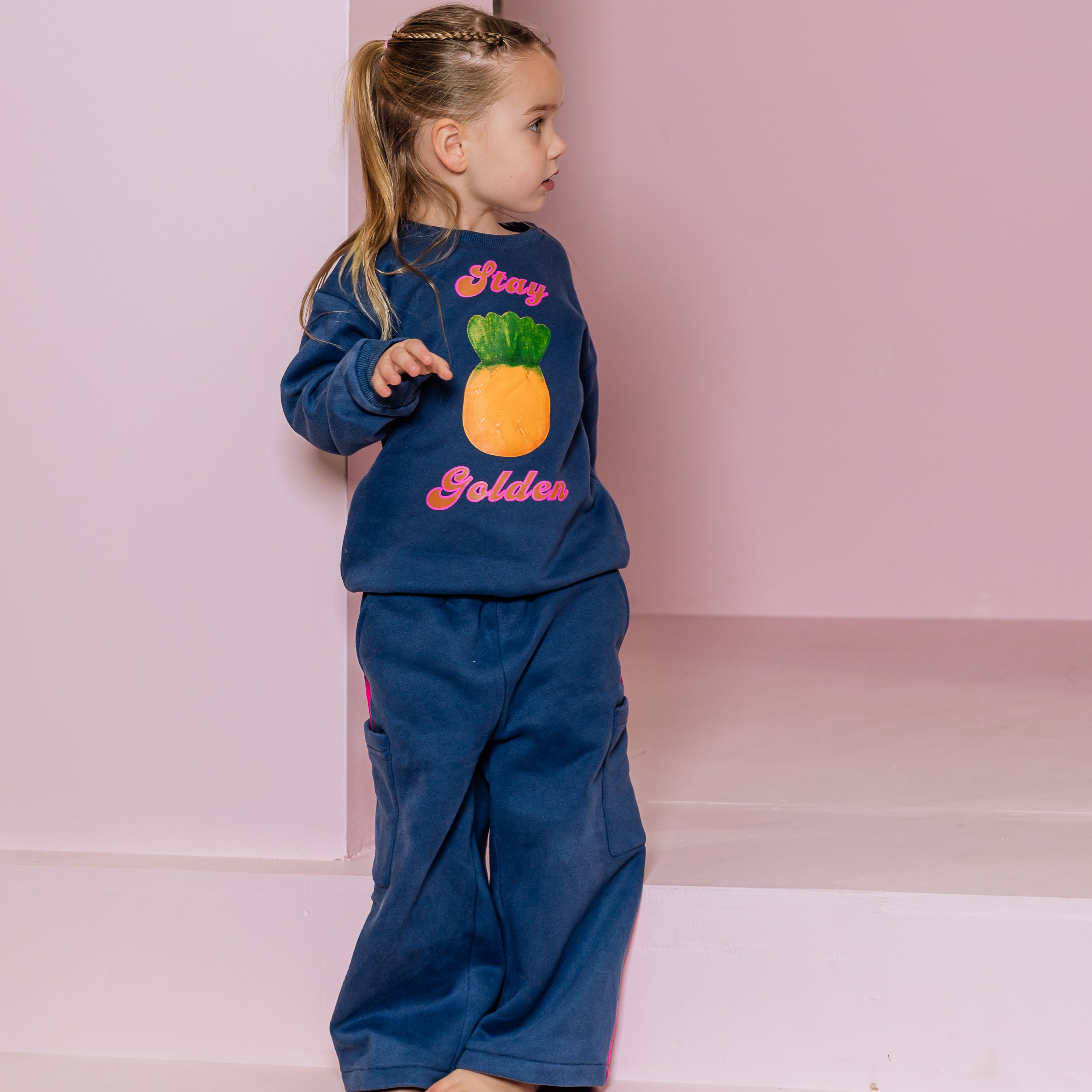 Child wearing a navy blue outfit with pineapple lolly design and 'Stay Golden' text on a pink background