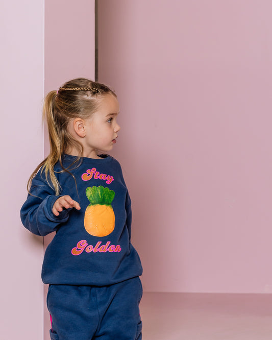 Child wearing a navy blue outfit with pineapple design and 'Stay Golden' text against a pink background