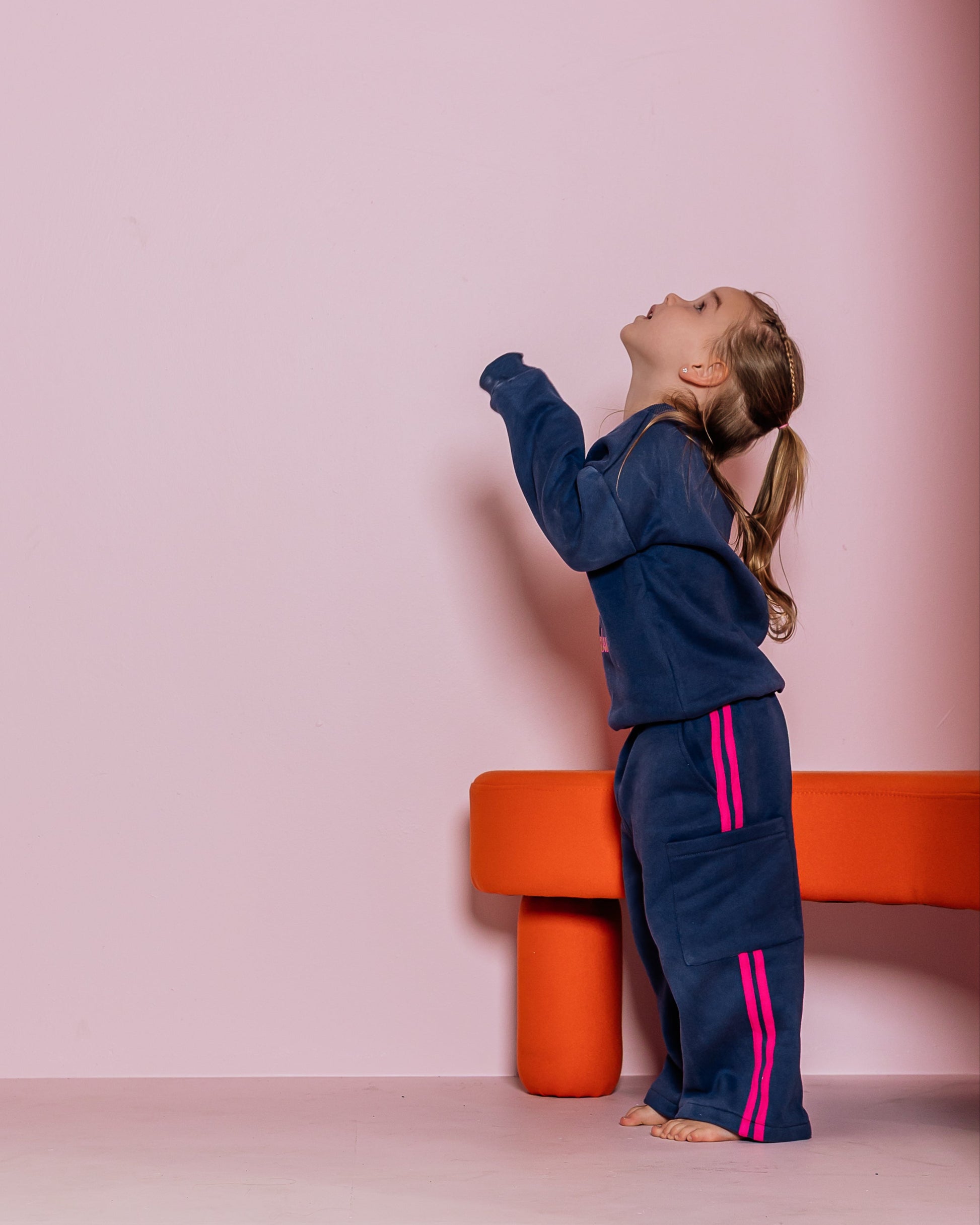 Child in a blue tracksuit with pink accents standing against a pink wall with an orange stool.