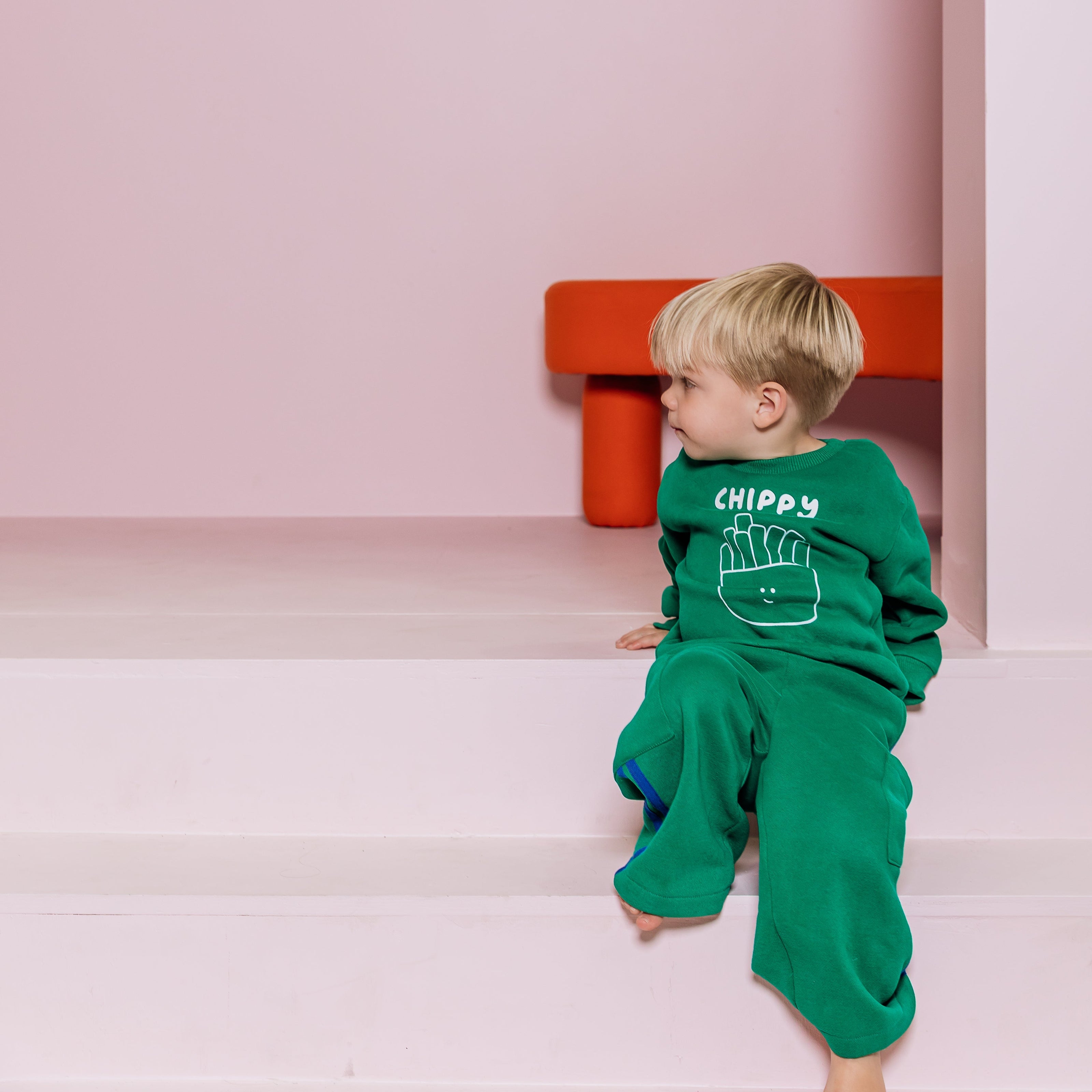 Child in green outfit with 'chippy thief' text and chips design sitting on a pink platform with a red object in the background