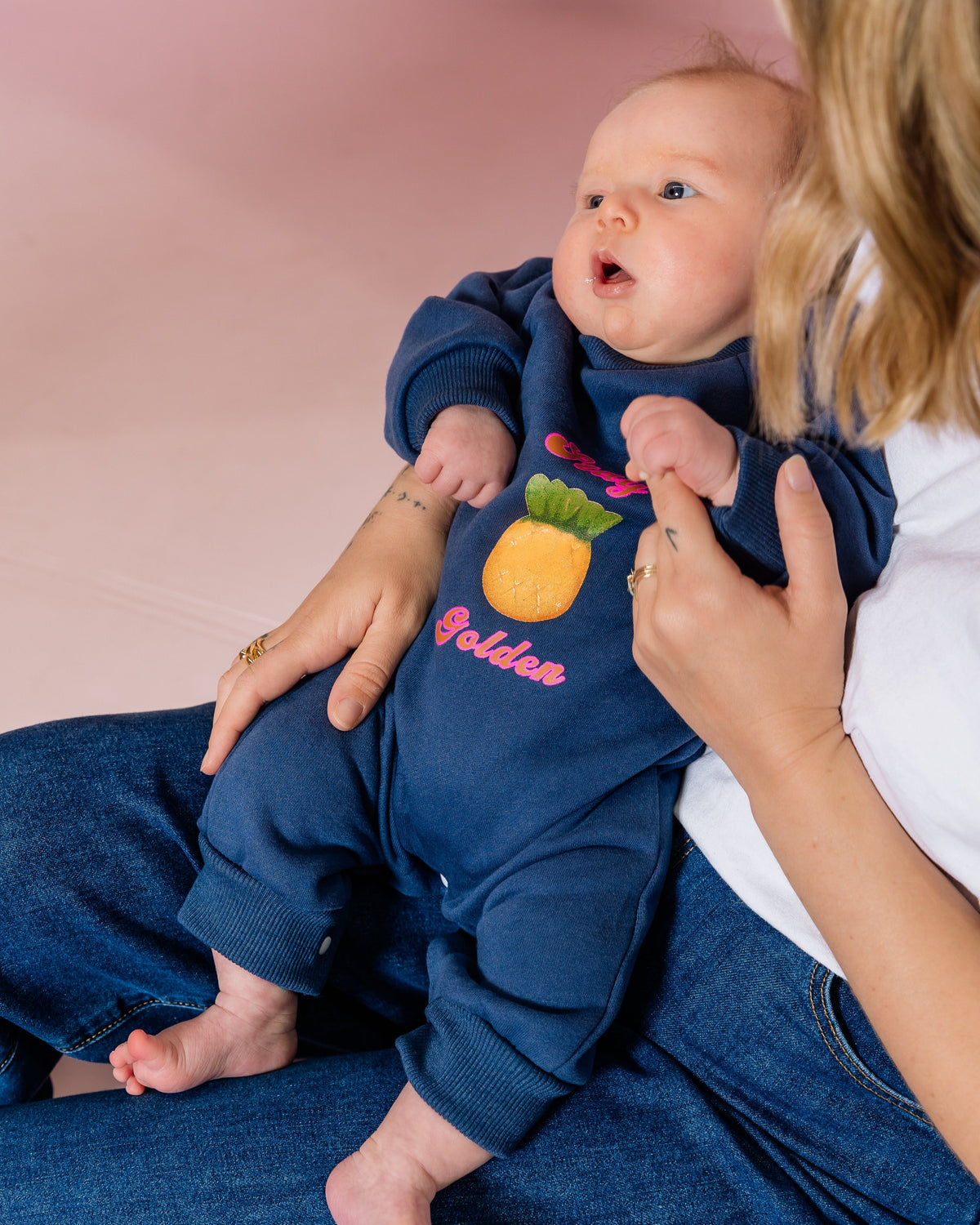 Baby in a navy blue outfit with a pineapple watercolour lolly design, sitting on their Mums lap against a pink background.