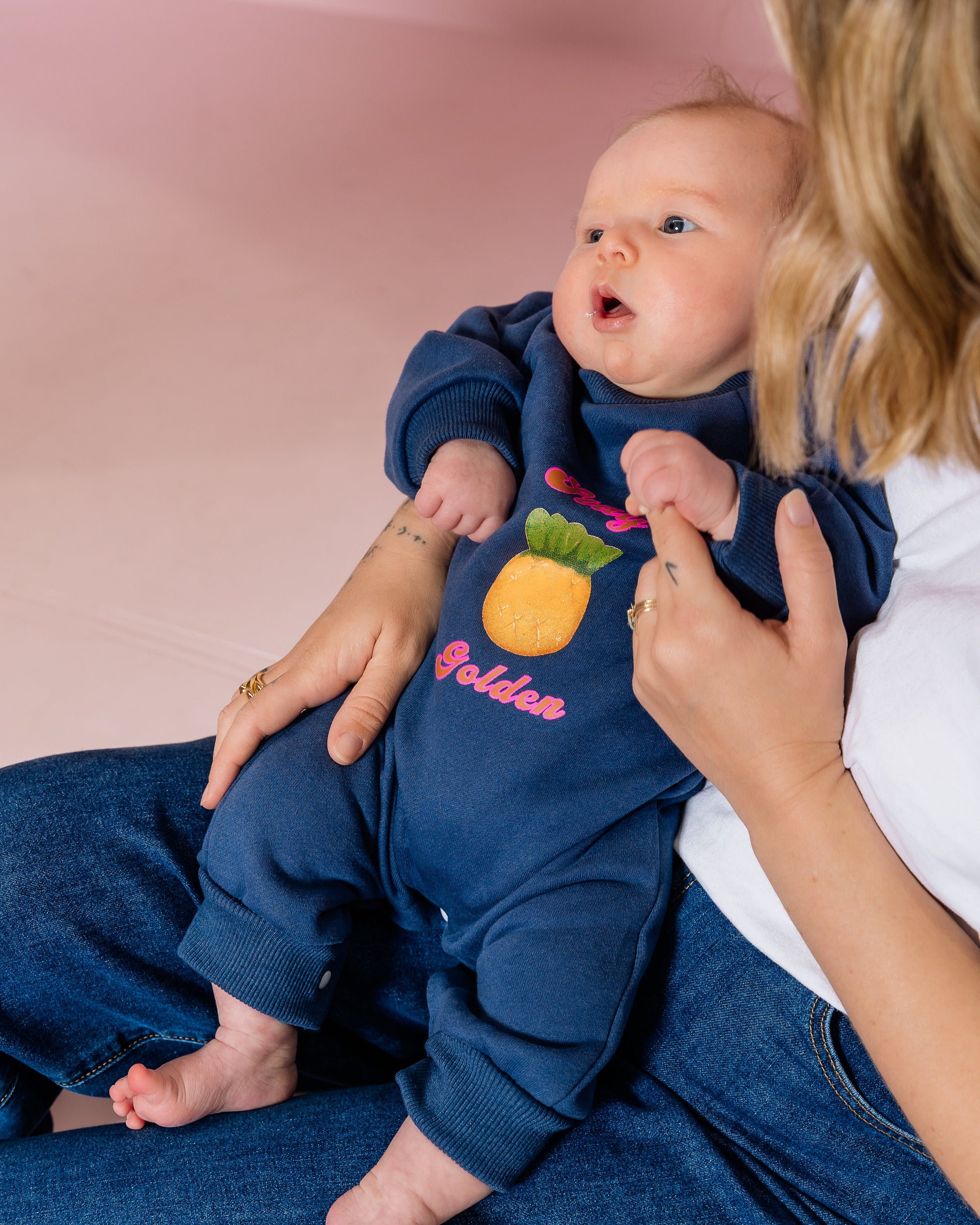 Baby in a navy blue outfit with a pineapple design, sitting on a person's lap against a pink background.
