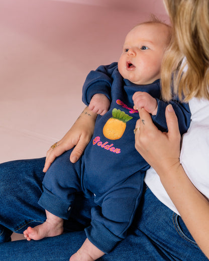 Baby in a navy blue outfit with a pineapple design, sitting on a person's lap against a pink background.
