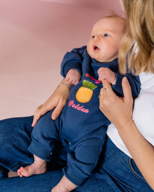 Baby in a navy blue outfit with a pineapple design, sitting on a person's lap against a pink background.