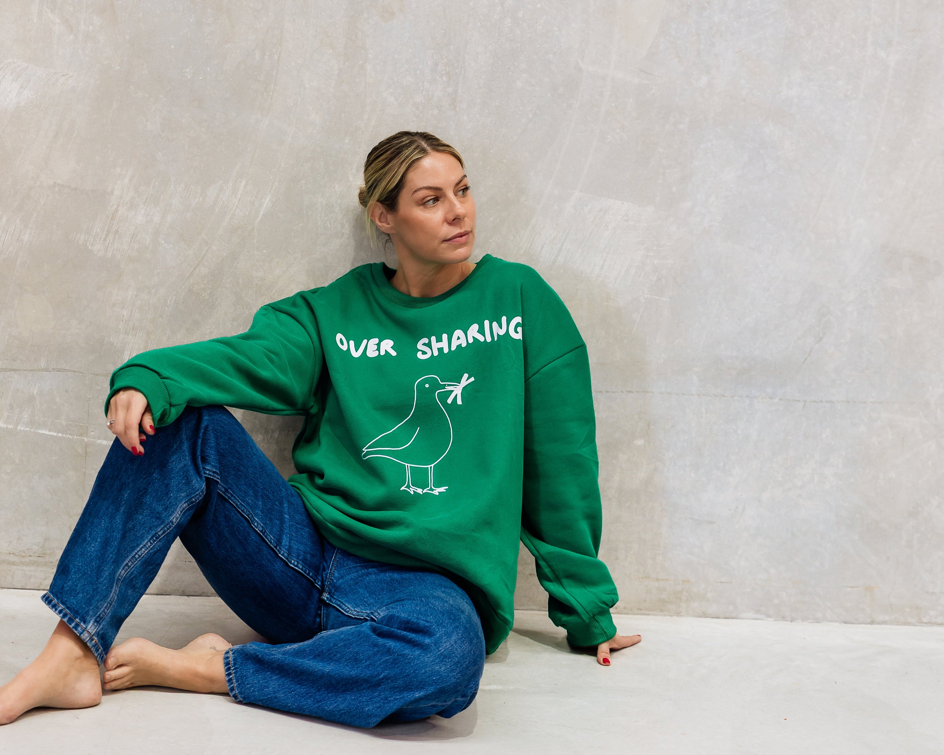 Person wearing a green sweatshirt with text "over sharing' and a graphic, of seagull with chips in its mouth sitting against a plain wall.