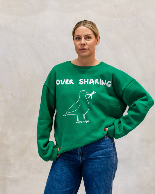 Person wearing a green sweatshirt with 'Over Sharing' text and a bird graphic against a plain background