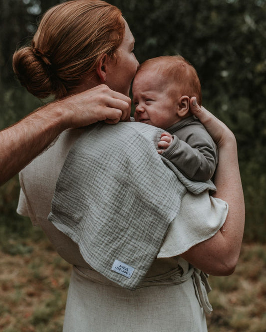 mother holds her baby over her shoulder with a warm brown slate burp cloth over the same shoulder. A mans arm helps her by adjusting the burp cloth to be under the babys head.
