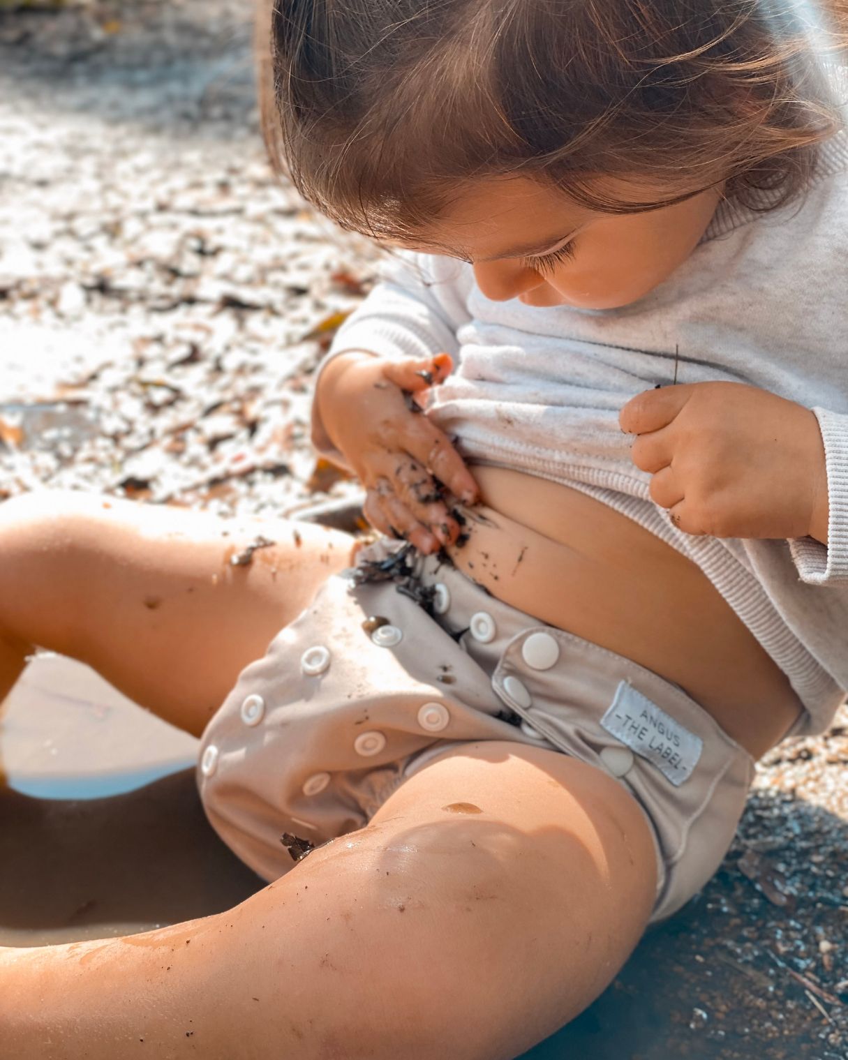 Toddler sits rubbing mud on her belly wearing a tan modern cloth nappy and grey shirt