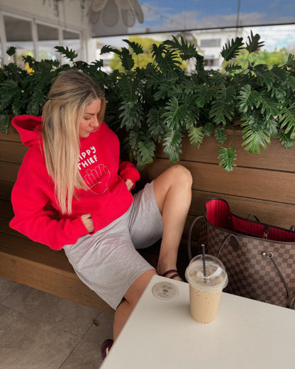 A woman sits on a wooden bench at an outdoor cafe, wearing a bright red hoodie with 'CHPPY THIEF' printed in white on the chest, paired with a light gray sweatshirt and shorts. A brown patterned handbag and an iced coffee are next to her.