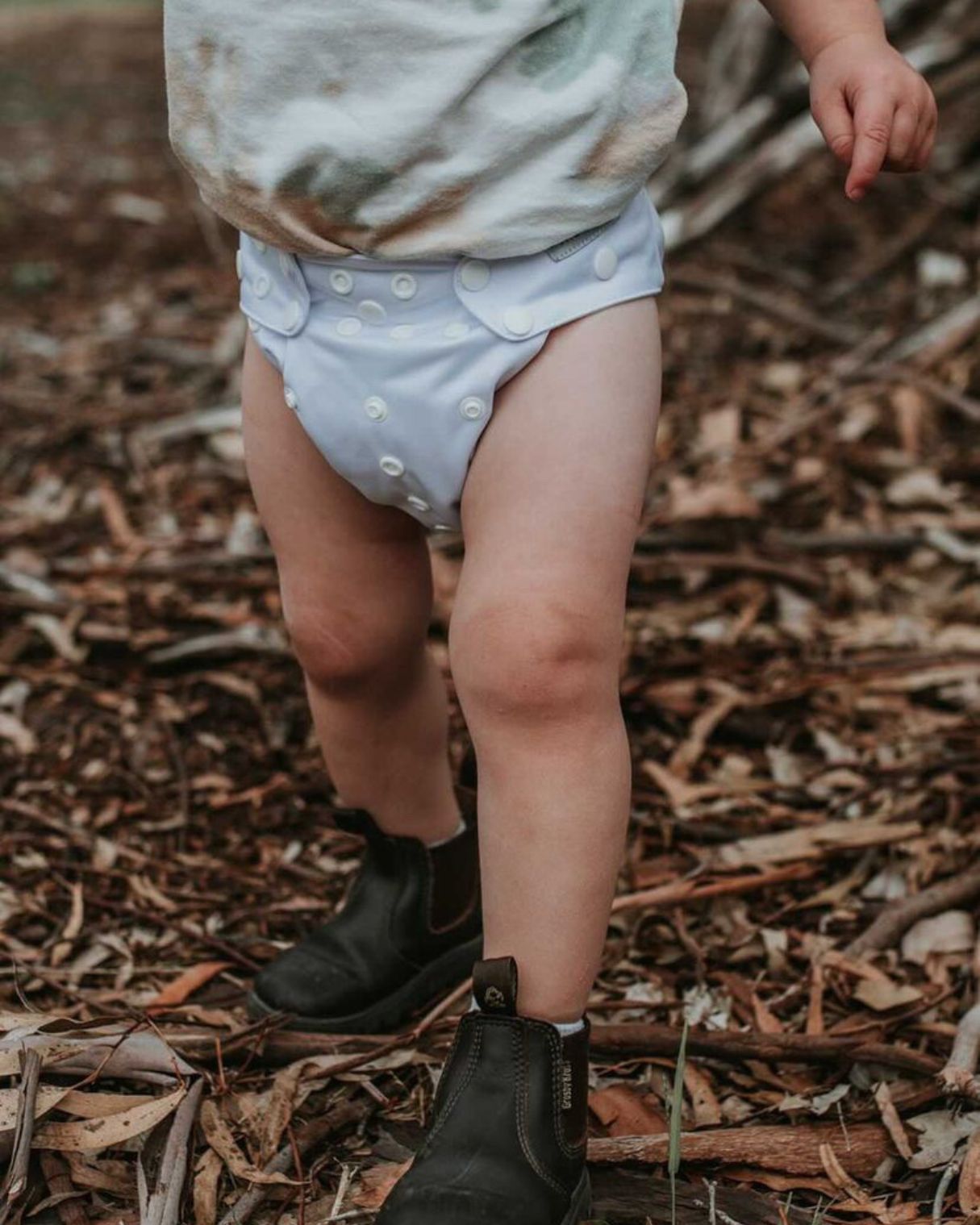 Toddler walks in his grey modern cloth nappy in some dead leaves and sticks.