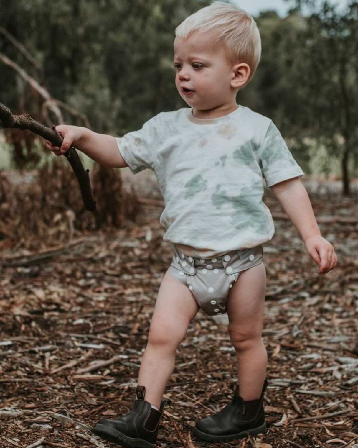 Toddler walks through the sticks wearing a tan modern cloth nappy holding a stick in the air