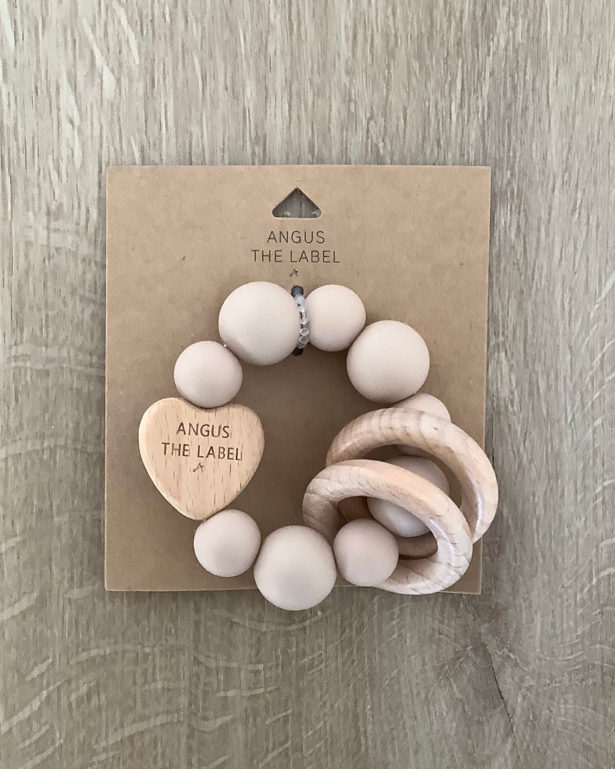 Taupe silicone beaded teething ring on a brown cardboard packaging sits on a wooden background. The teething ring has a beechwood heart and two beechwood rings. The heart has Angus The Label lasered onto it.