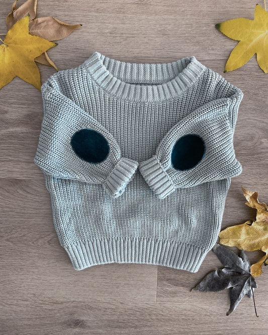 Minty green knit toddlers sweater with moss green coloured elbow patches. Background is wooden floorboards and Autumn leaves