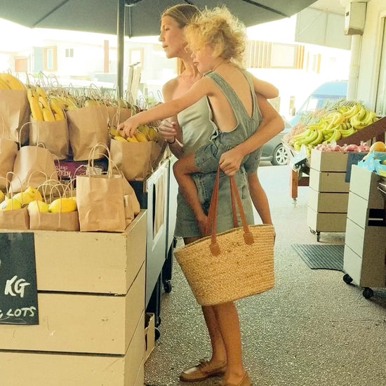 A mother shopping at an outdoor market with her child, carrying a stylish straw tote bag filled with fresh produce, showcasing Angus The Label's practical and fashionable accessories.