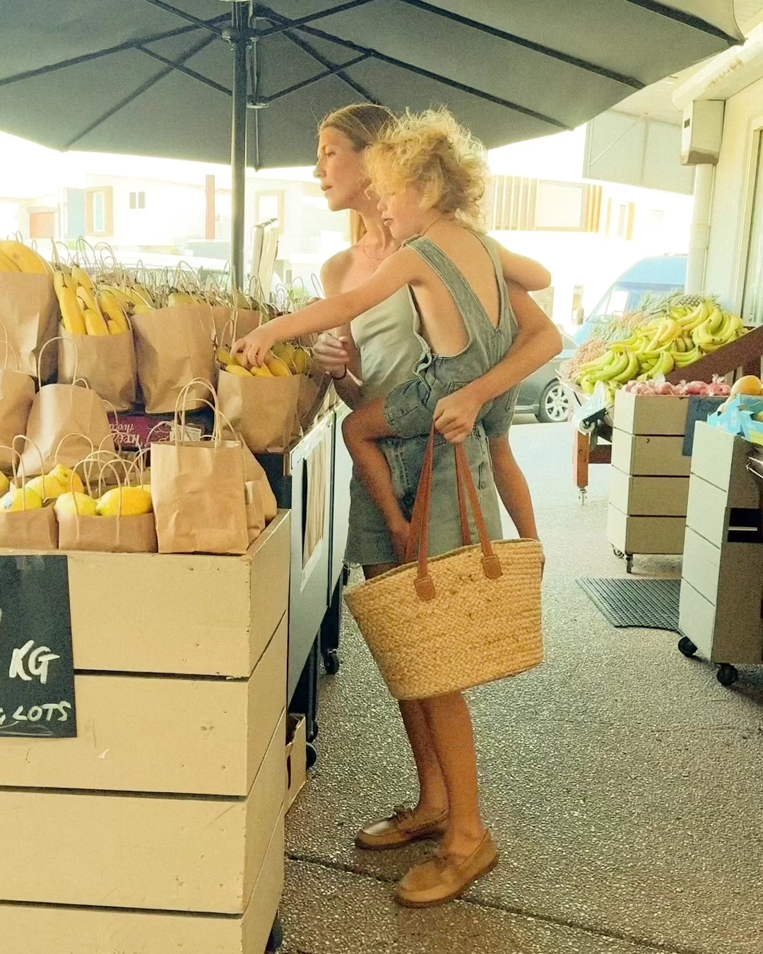 A mother shopping at an outdoor market with her child, carrying a stylish straw tote bag filled with fresh produce, showcasing Angus The Label's practical and fashionable accessories.