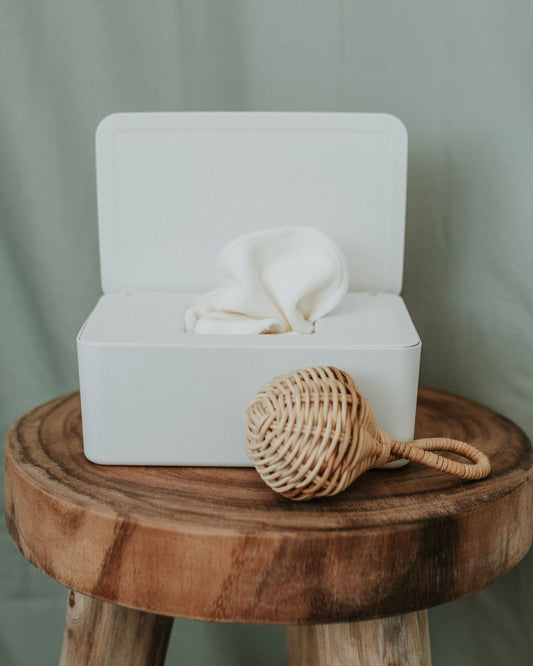 Cream wipe container with bamboo washers in it. Sitton on a wooden side table with rattan baby rattle in front.